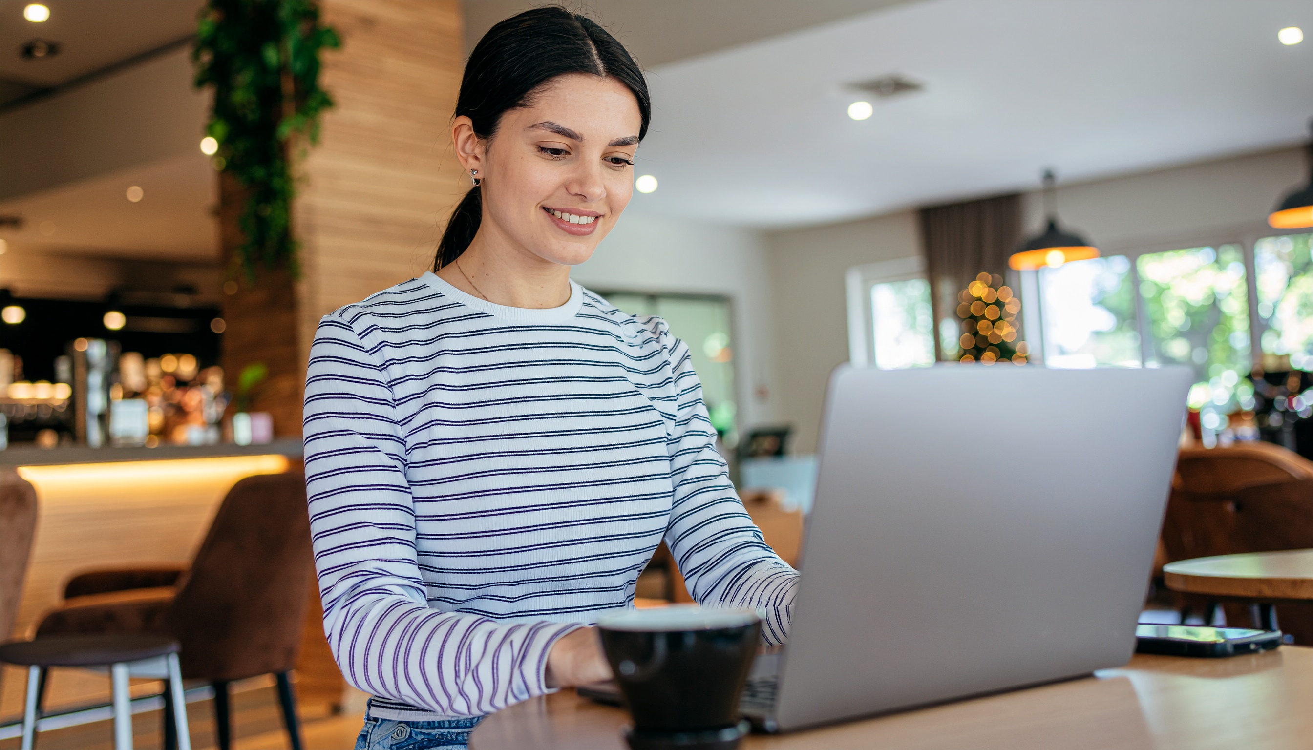 Mulher sorrindo com camisa listrada usa laptop em um café moderno, luz suave, detalhes em madeira e decoração verde, criando um ambiente acolhedor e contemporâneo.